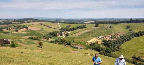 Two people walking uphill in a lush, green rural landscape with rolling hills, scattered farms, and small ponds. The background features distant forests and a partly cloudy blue sky.